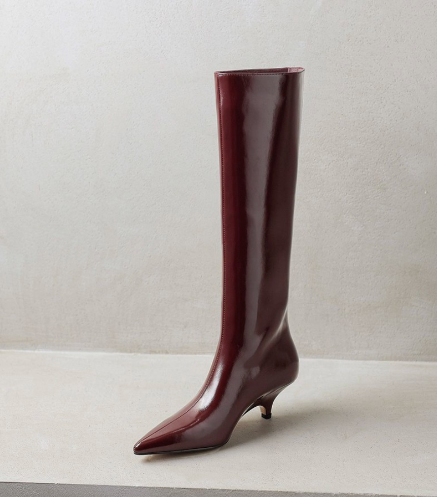 Mescini burgundy kitten heel knee high boots, one boot standing on a pale set, US women's sizes