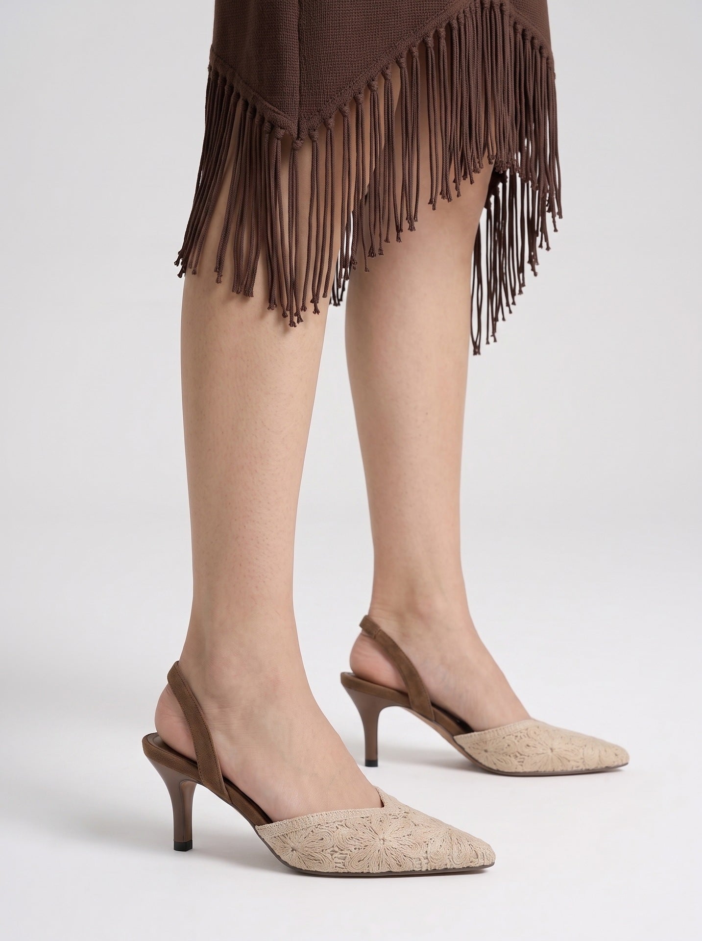 Mescini brown crochet pointed toe slingback heels, worn standing with a fringed hem behind, US women's sizes